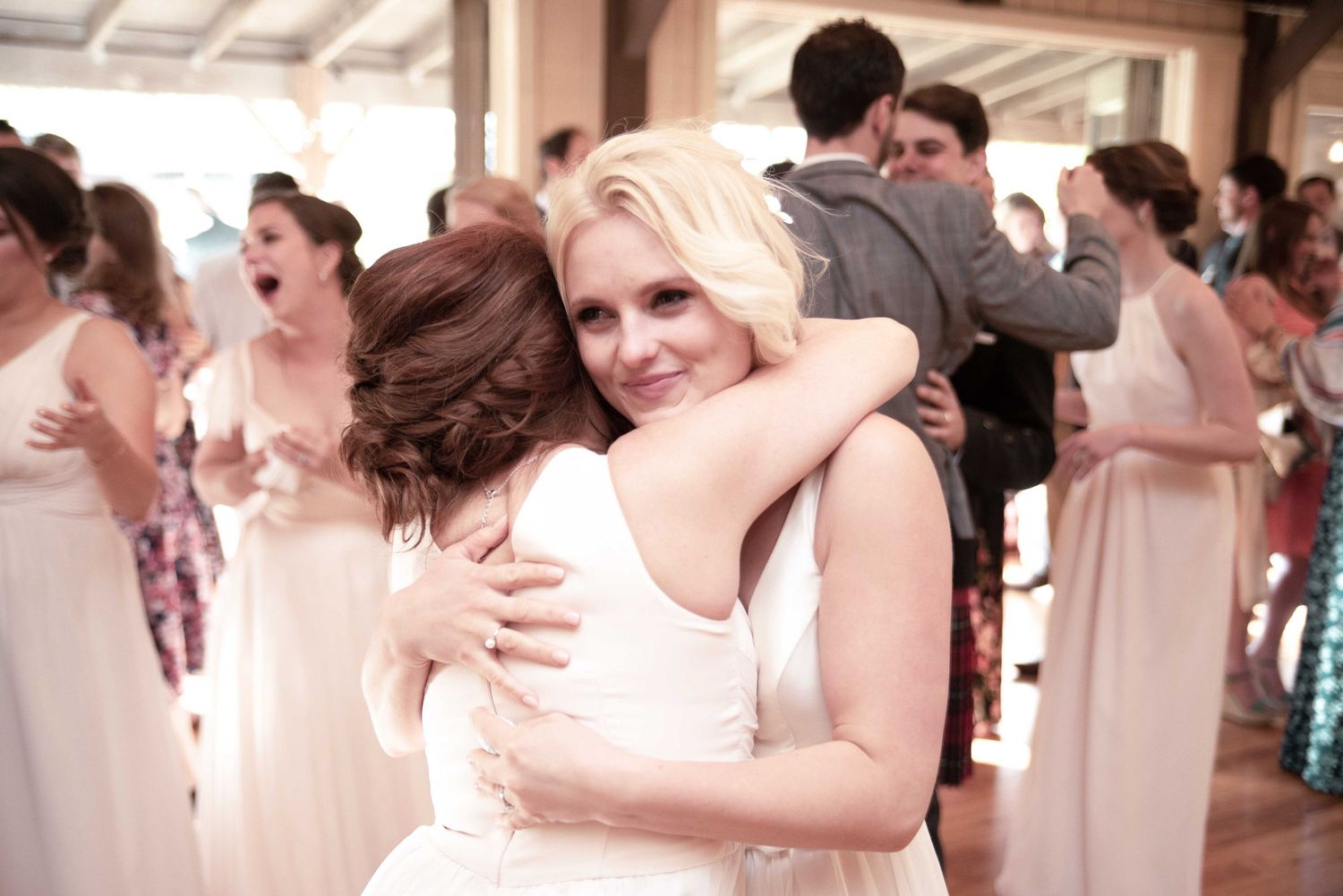 Two women in white dresses share a warm hug at a wedding reception with guests dancing in the background.