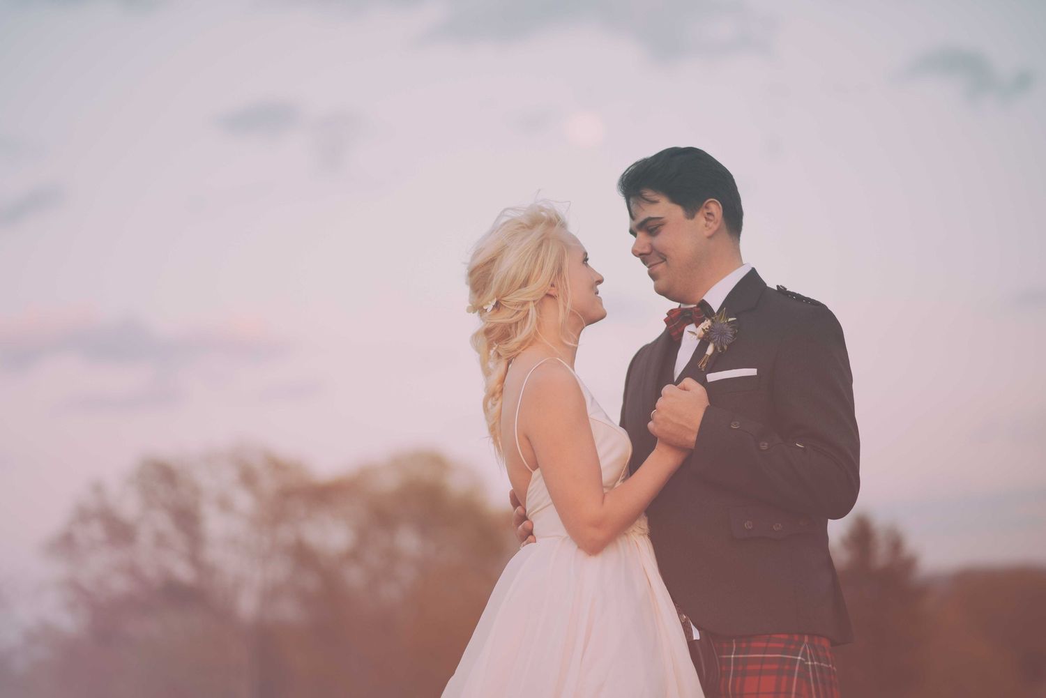 Bride in white dress and groom in dark suit with red tartan kilt embrace outdoors at sunset.