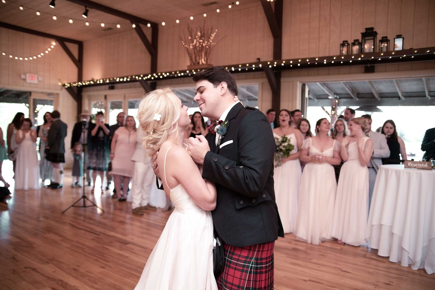 Bride and groom share first dance in rustic barn venue, groom wearing traditional Scottish tartan kilt.