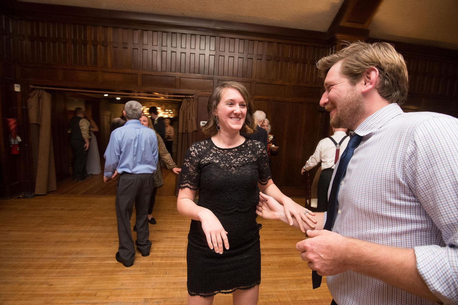 A man and woman smile and hold hands while dancing at an elegant wood-paneled ballroom event.
