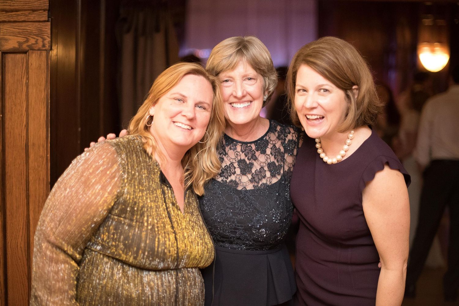 Three women smiling and posing together at a formal evening event in an elegant venue.