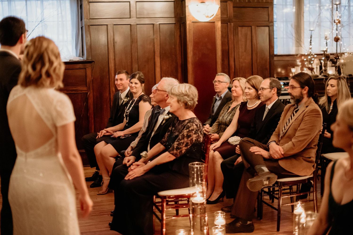Wedding guests seated in wooden chairs watching ceremony in elegant wood-paneled room with candlelight.