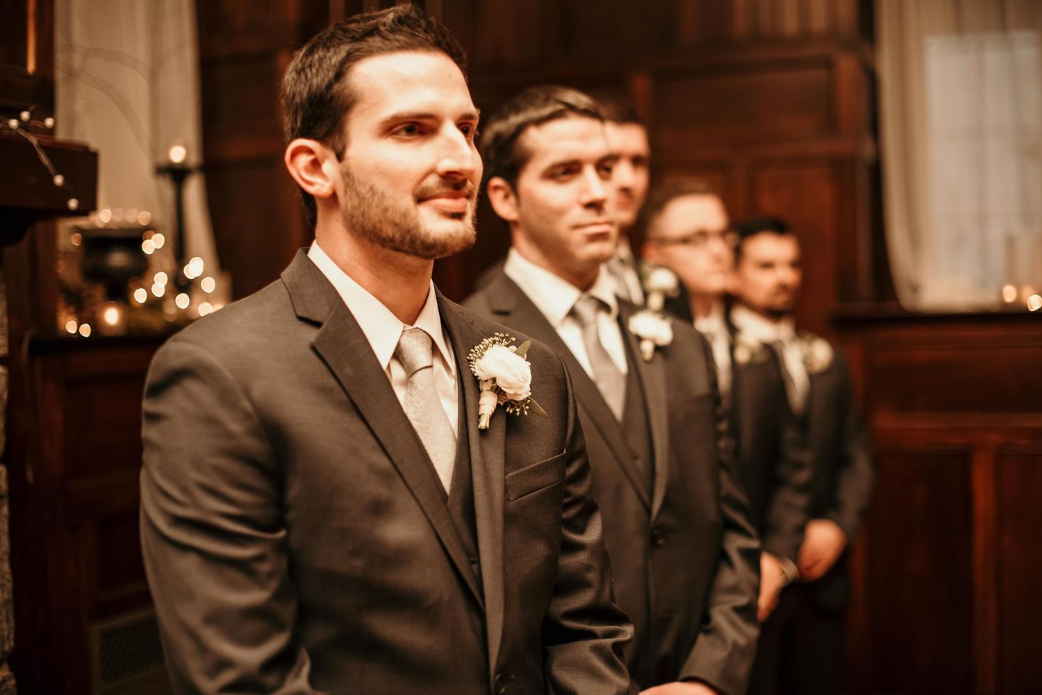 Groomsmen in gray suits with boutonnieres standing in a row at a wedding ceremony indoors.