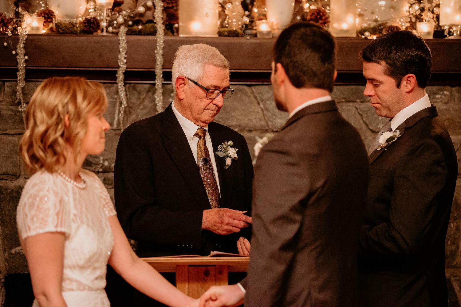 Officiant reads vows during intimate indoor wedding ceremony by stone fireplace with candles and autumn decor.