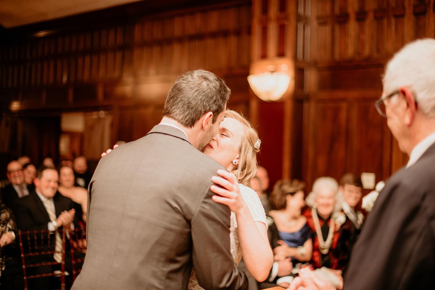 Bride and groom share their first kiss at wedding ceremony with guests applauding in elegant wood-paneled venue.