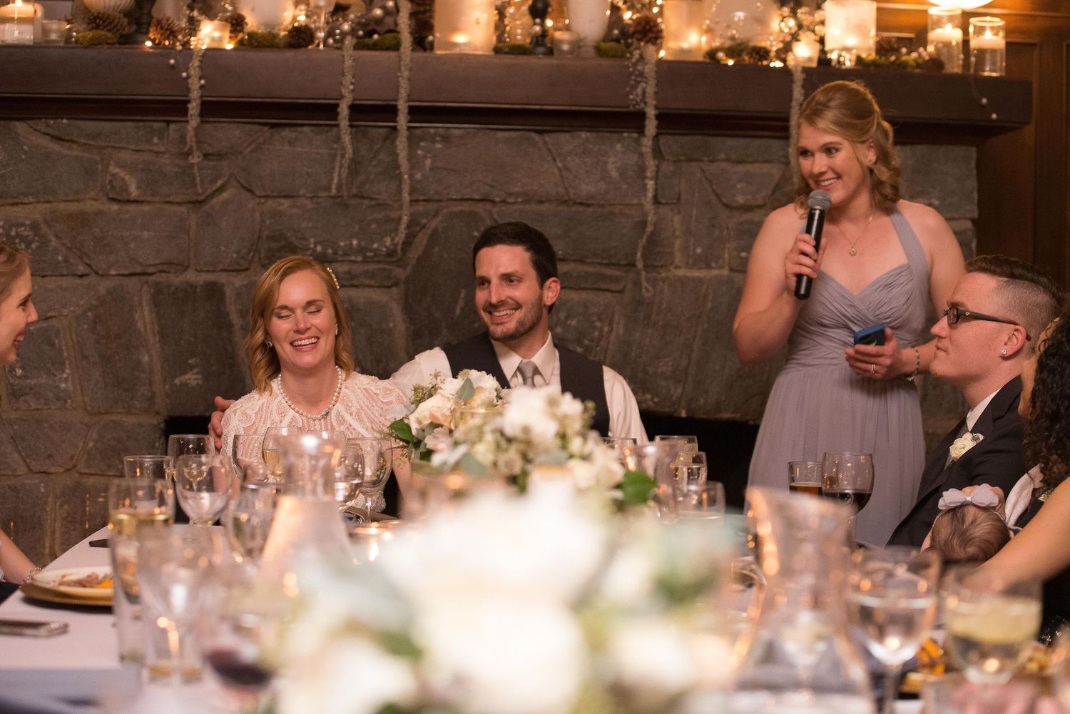 Wedding reception toast with bridesmaid speaking at elegant candlelit venue with stone fireplace backdrop.