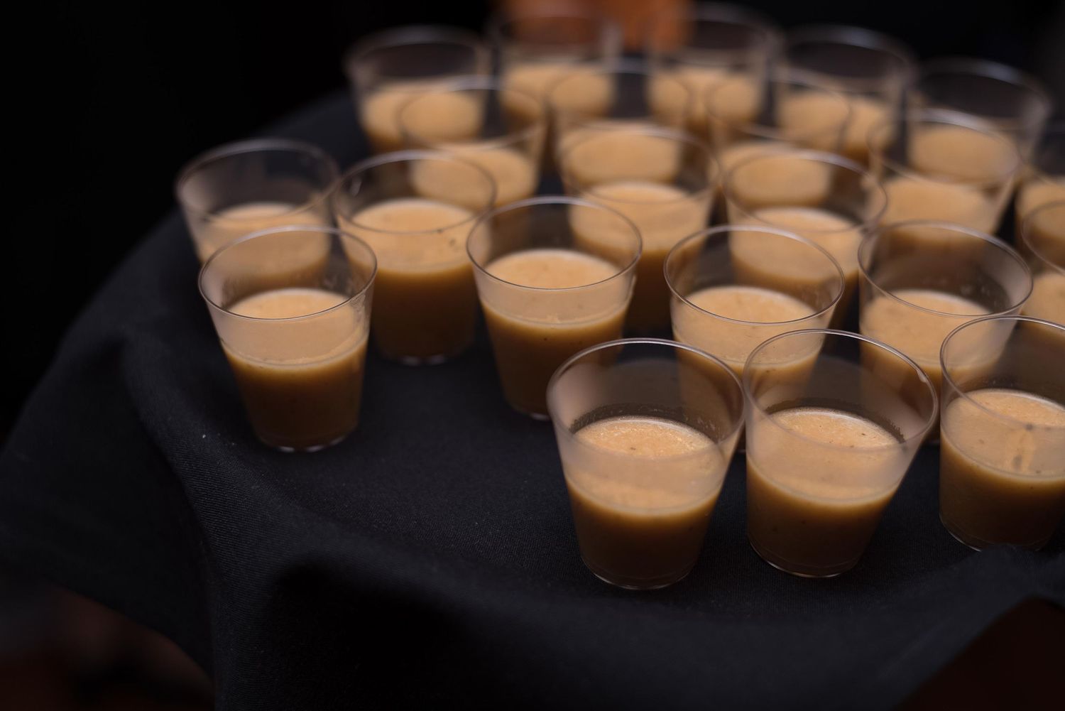 Rows of small plastic shot glasses filled with creamy coffee liqueur served on a dark cloth-covered tray.