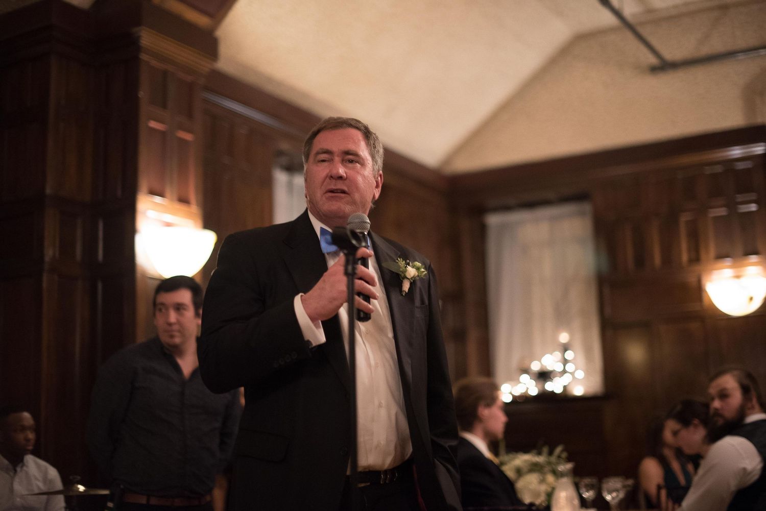 Man in suit giving speech with microphone at elegant wood-paneled banquet hall event.