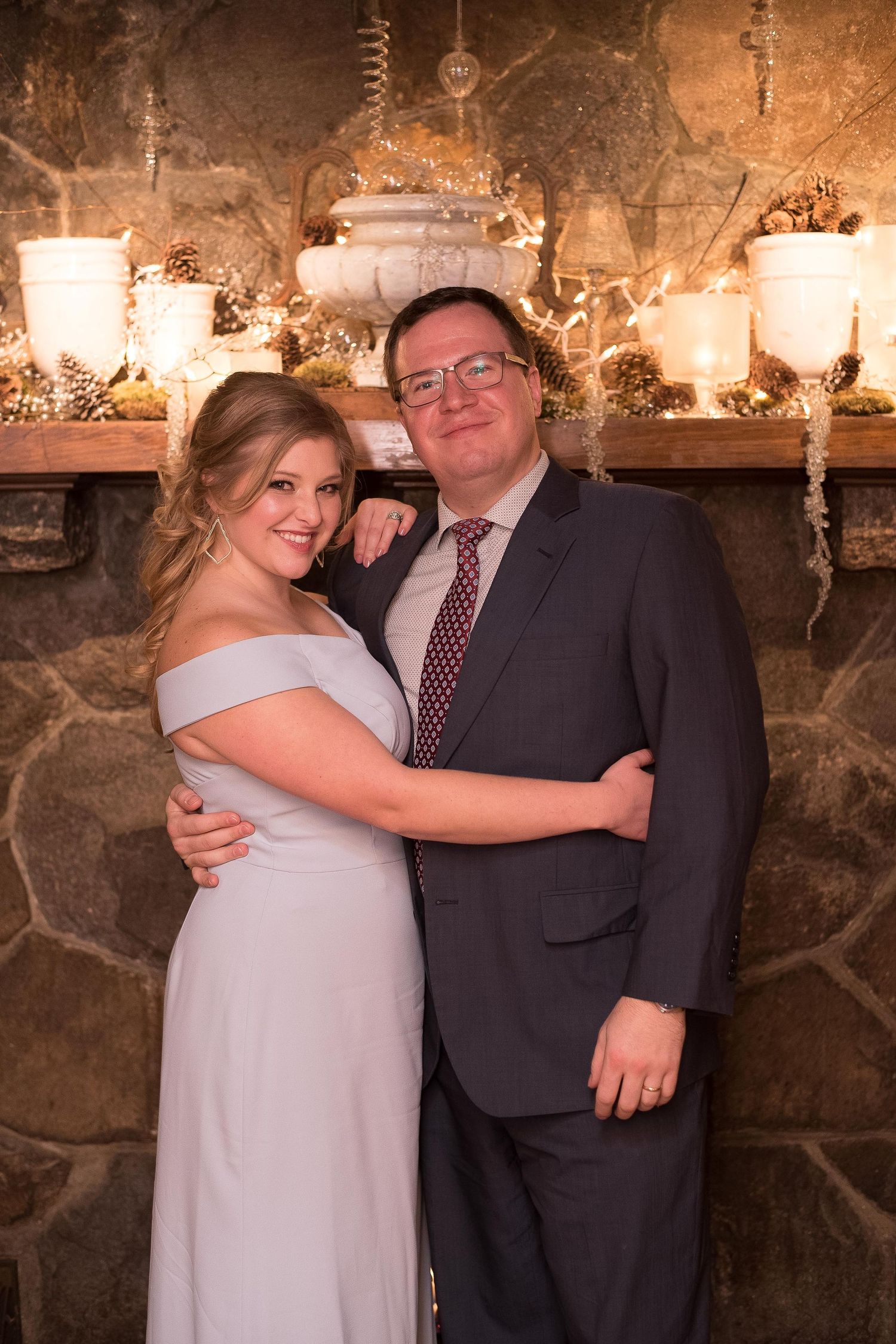 Couple posing together in formal attire in front of a stone fireplace decorated with candles and pinecones.