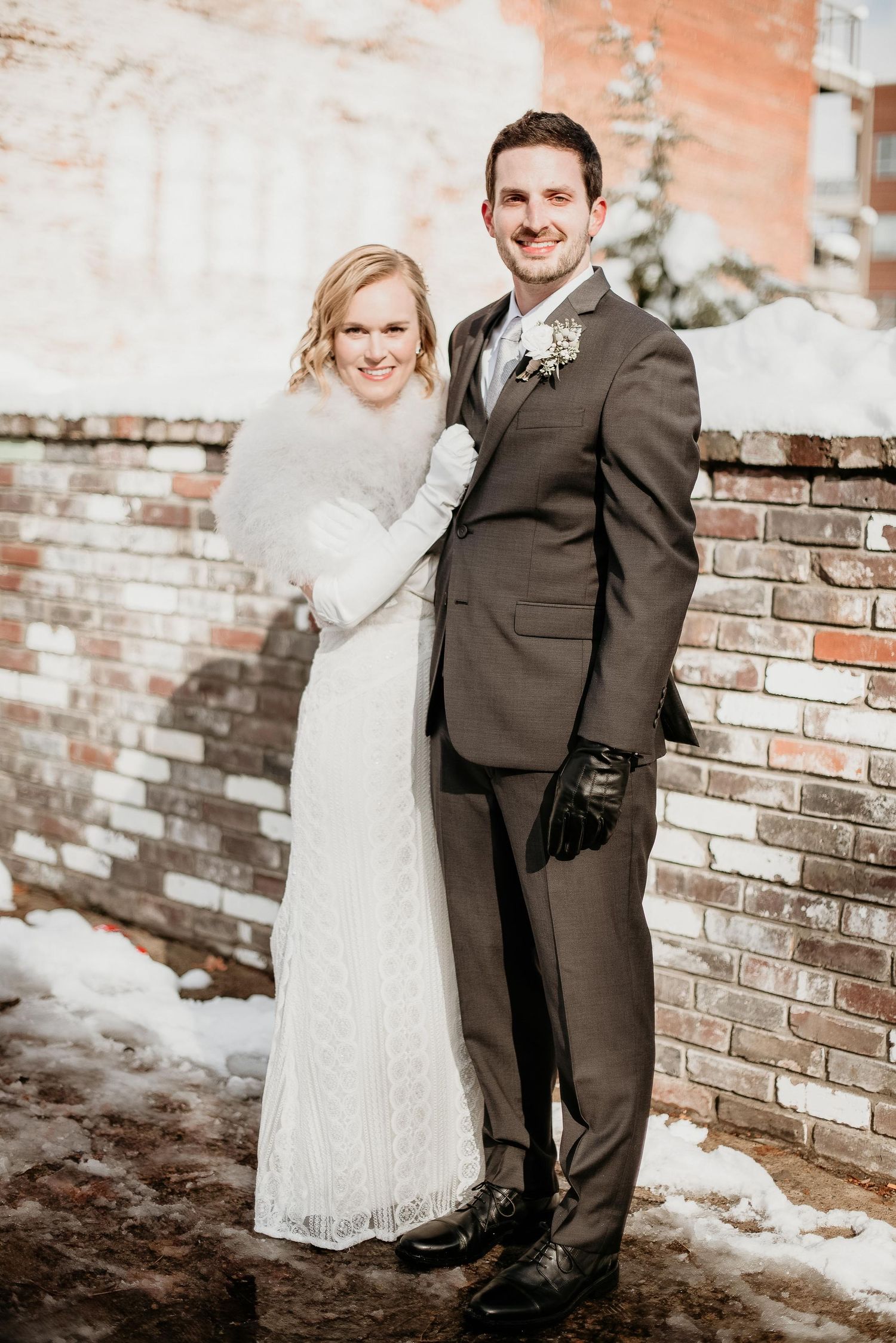 Bride and groom pose together in winter wedding attire beside a brick wall dusted with snow.