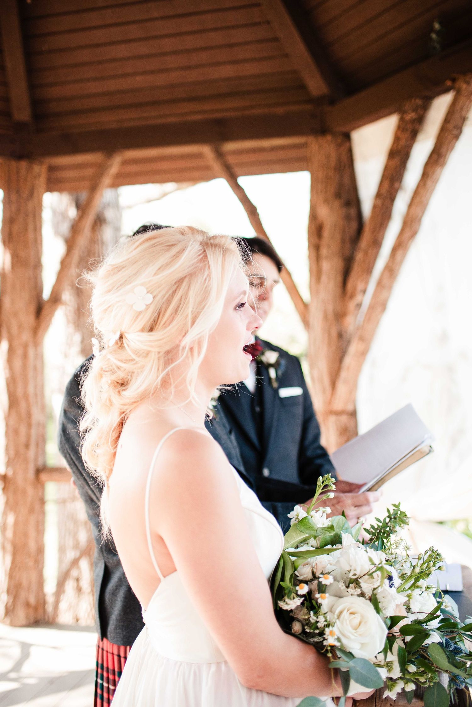 Blonde bride holding white floral bouquet reading vows under rustic wooden gazebo during outdoor wedding ceremony.