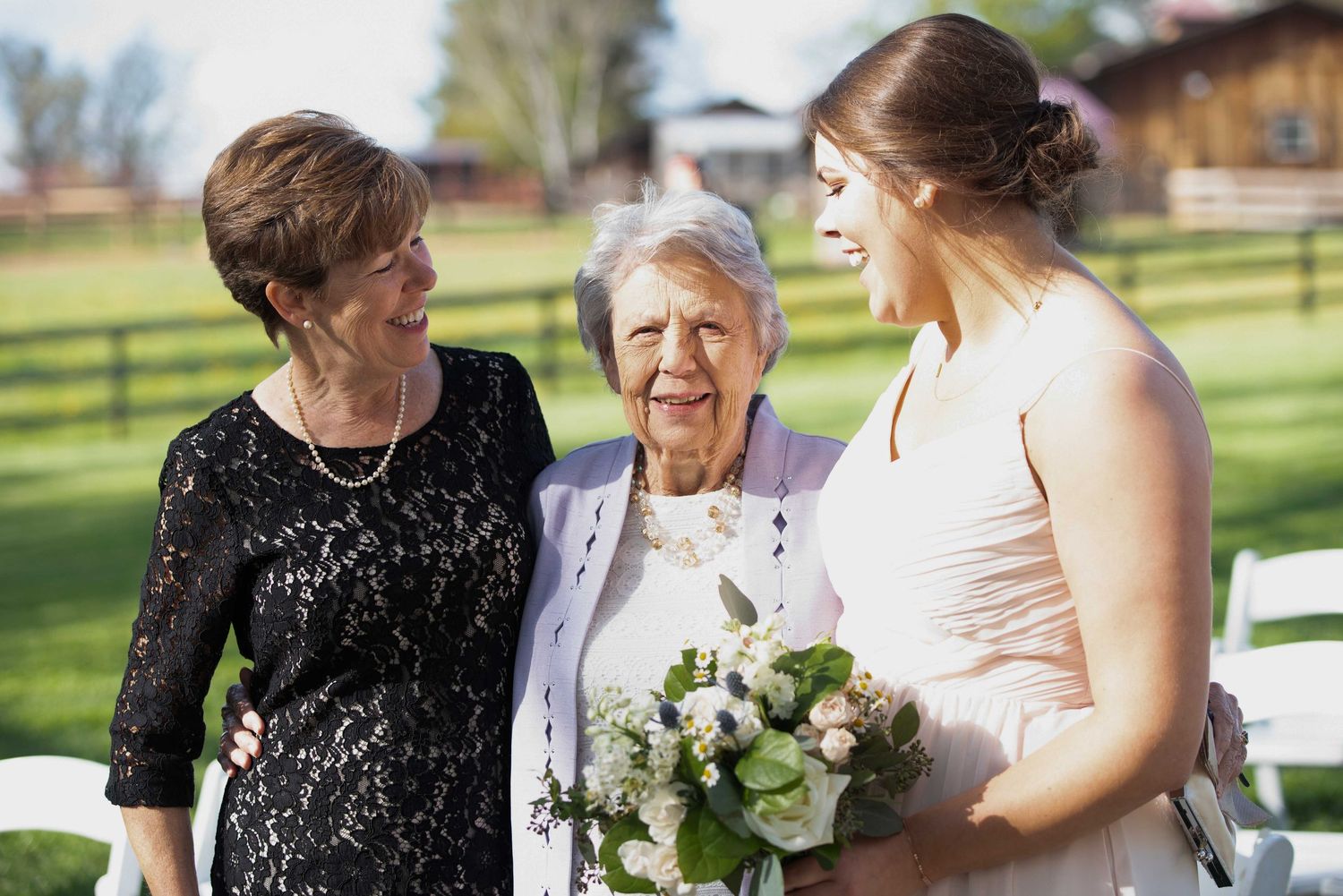 Bride holding bouquet smiles with two older women at outdoor wedding venue with green lawn.