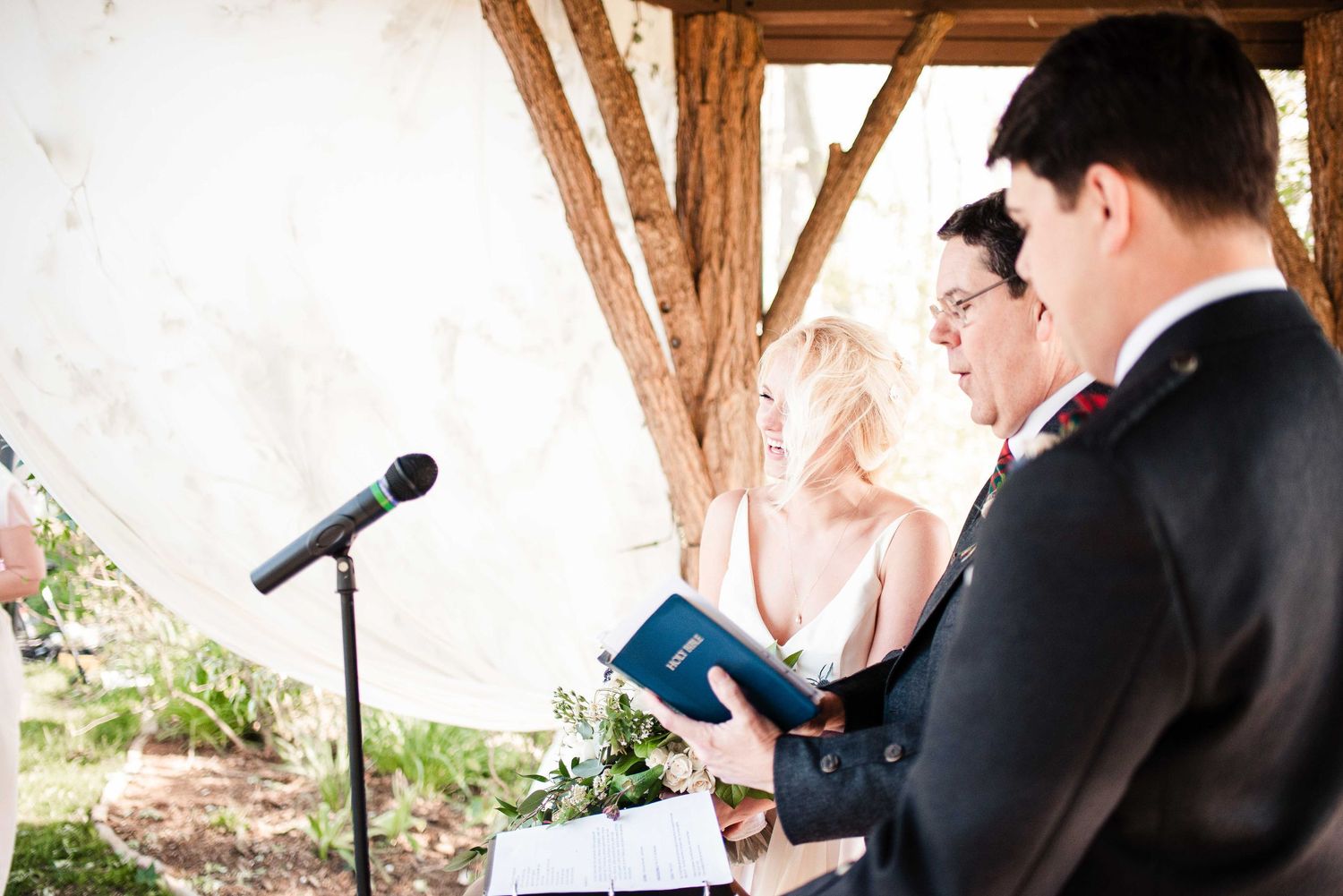 Officiant reads from blue book during outdoor wedding ceremony under rustic wooden structure with bride and groom.