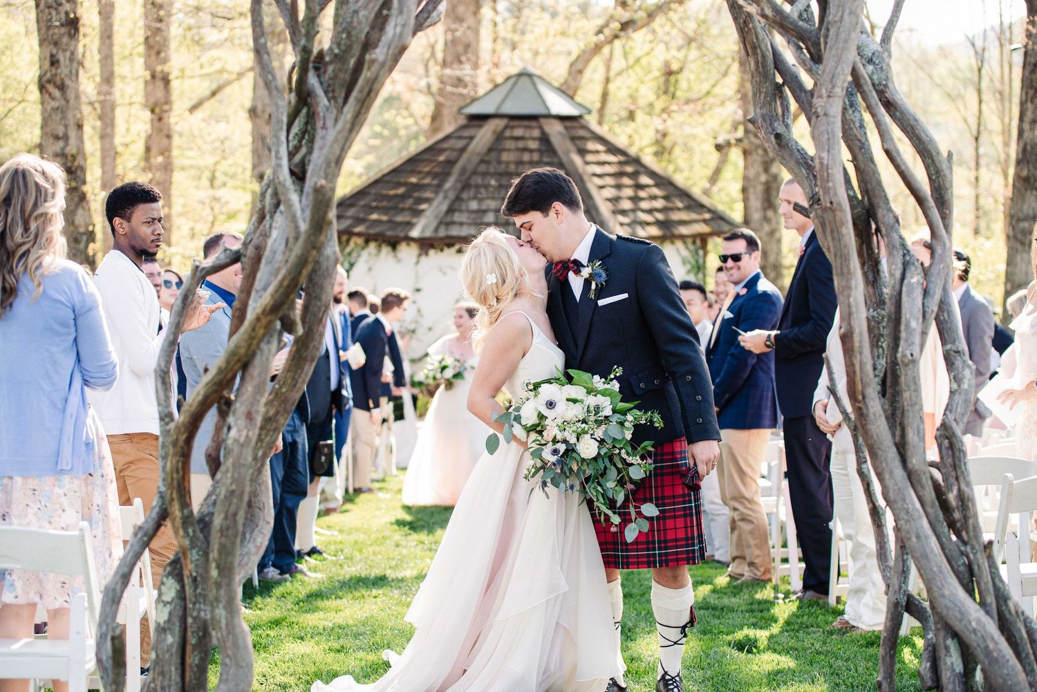 Bride and groom share a kiss at outdoor wedding ceremony, with guests in kilts and floral arch backdrop.