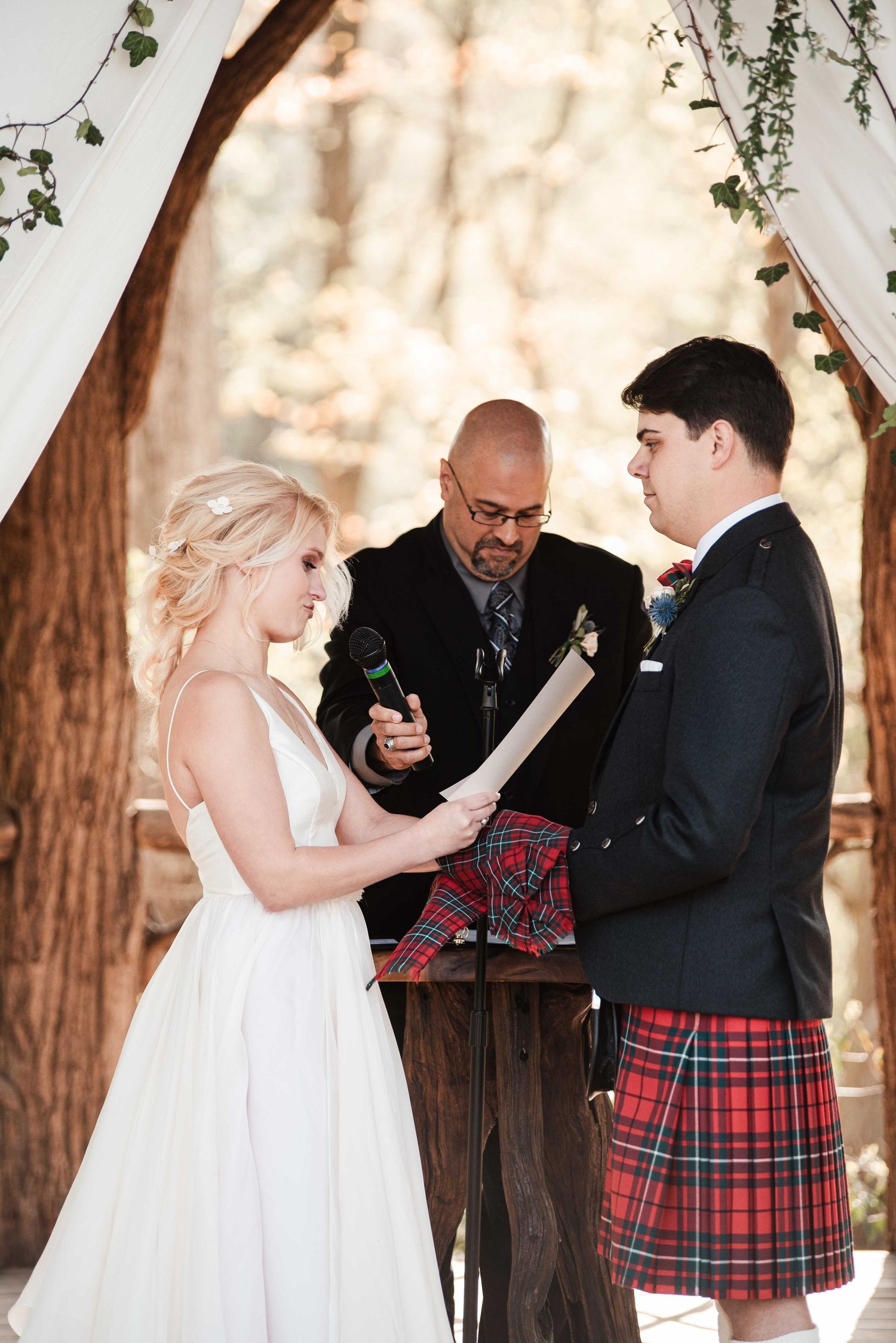Bride in white gown reads vows to groom in red tartan kilt at outdoor woodland wedding ceremony with officiant.