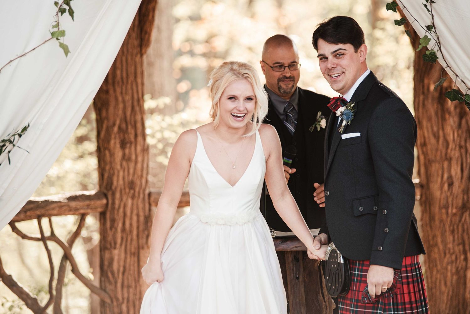 Smiling bride in white gown and groom in red tartan kilt celebrate after wedding ceremony outdoors.