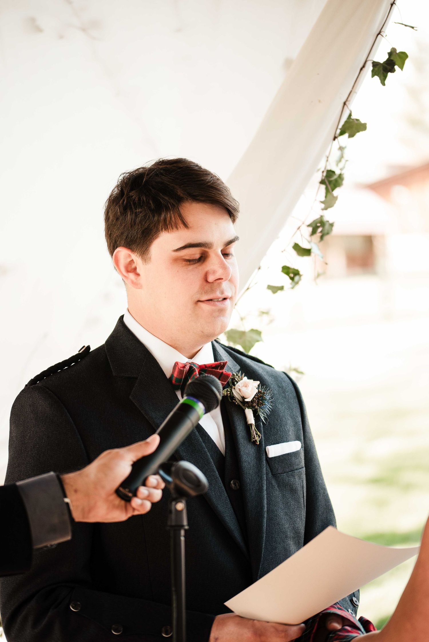 Groom in dark suit with red bow tie and boutonniere reads wedding vows at outdoor ceremony microphone.