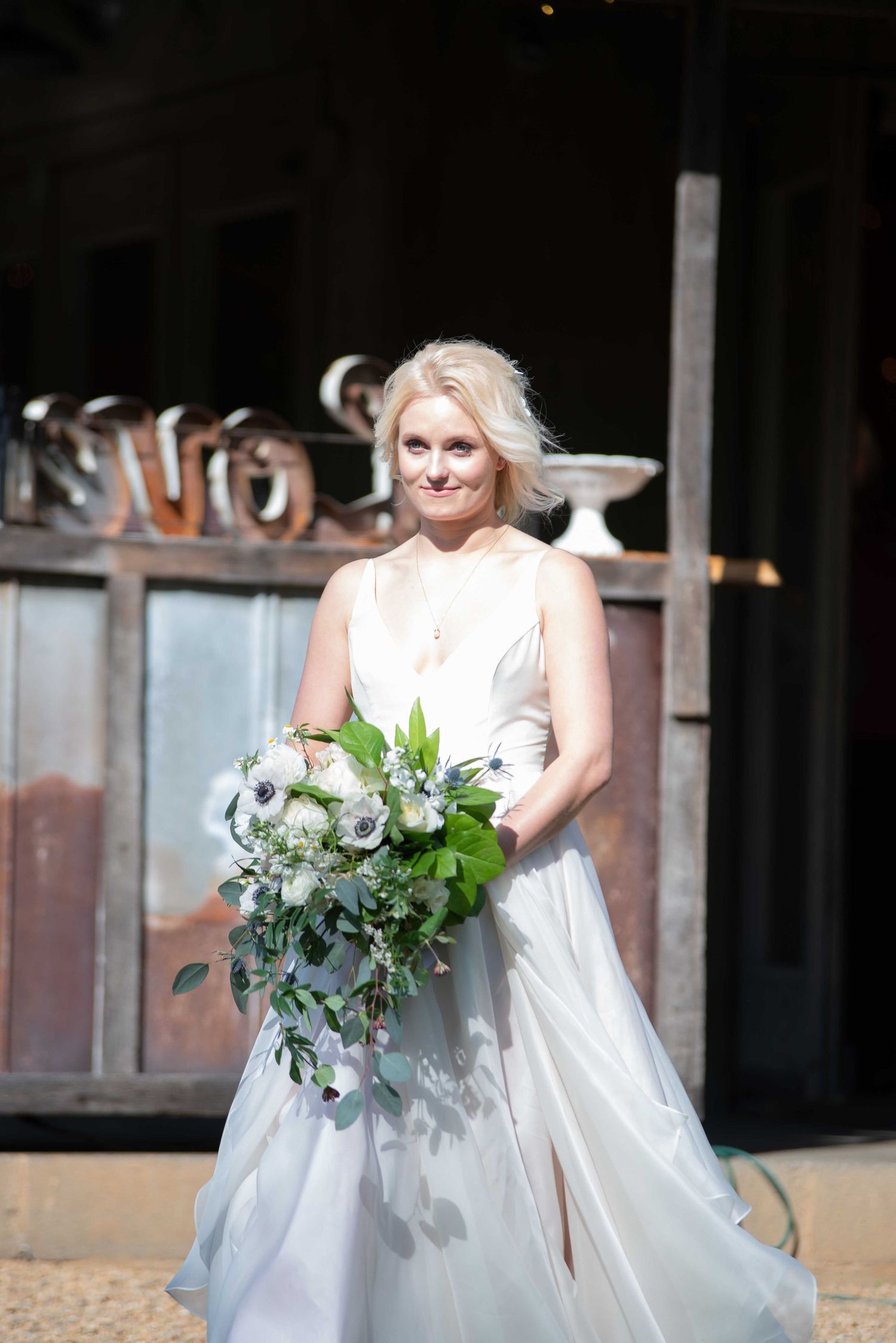 Blonde bride in white gown holding cascading greenery and white floral bouquet, posing outdoors at rustic venue.