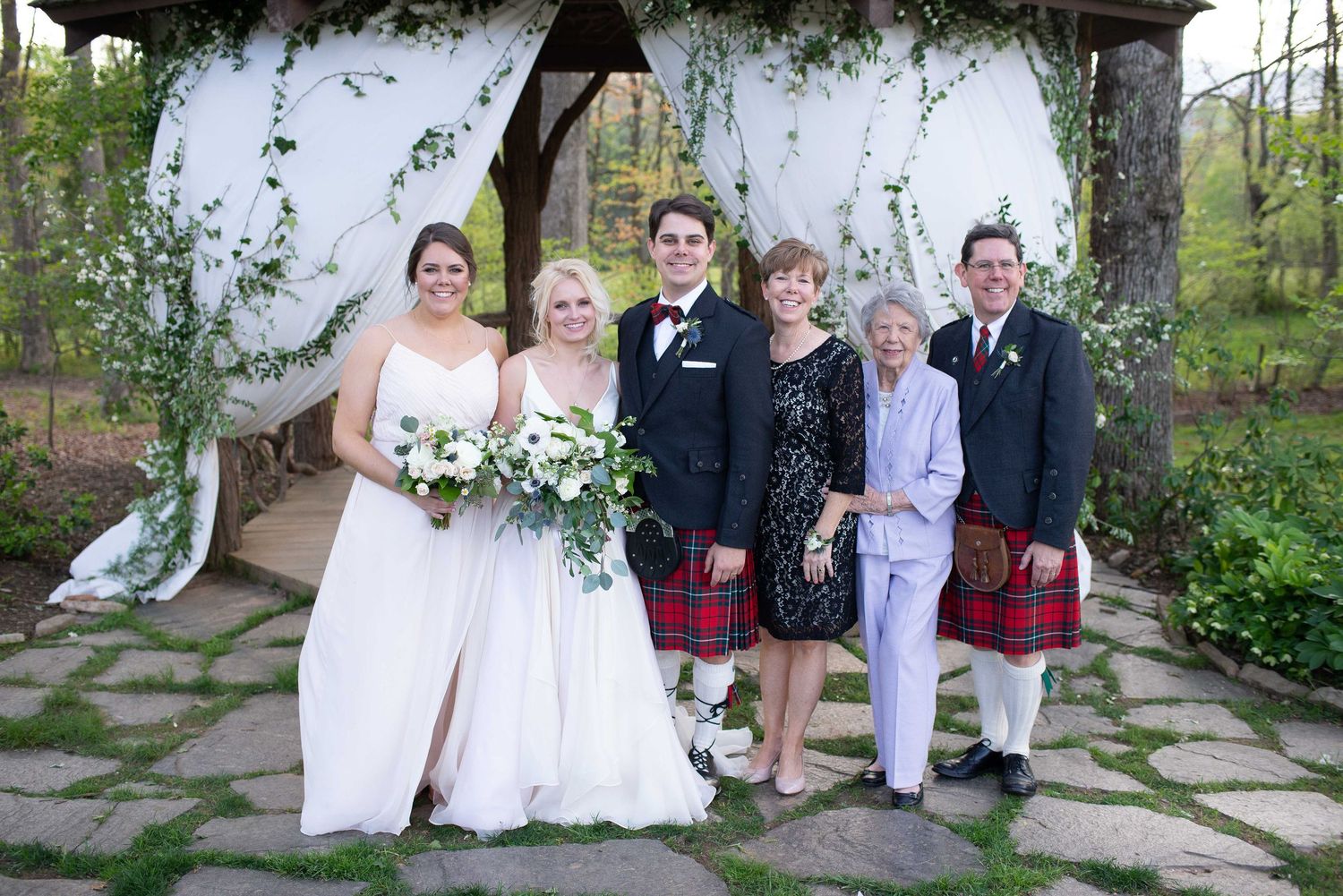 Wedding party posing under floral arch, two men in kilts, bride in white gown, bridesmaids in pink dresses.