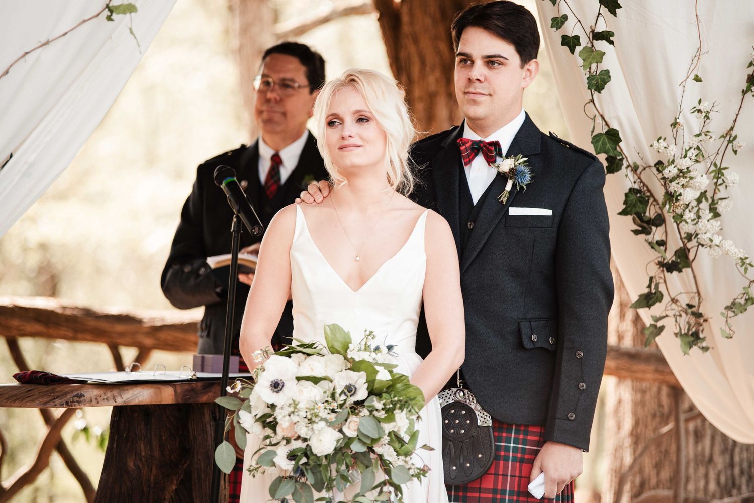 Bride holding floral bouquet stands with groom in Scottish kilt at outdoor wedding ceremony with officiant.