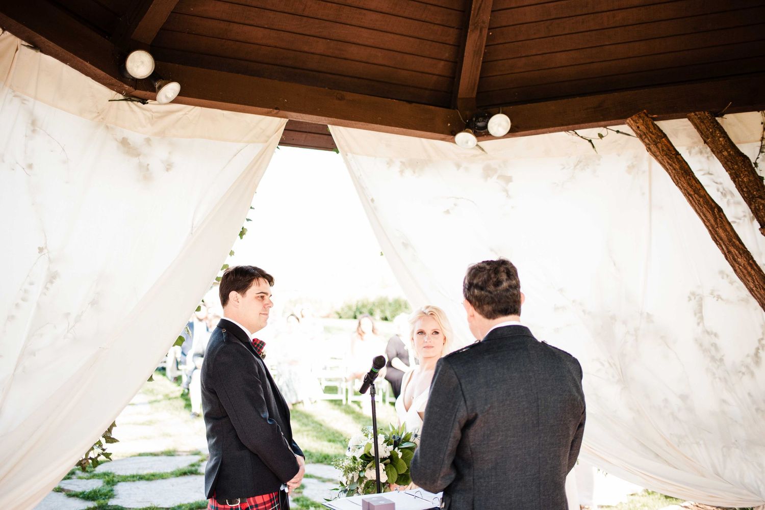 Outdoor wedding ceremony under a draped gazebo with string lights, officiant facing couple.