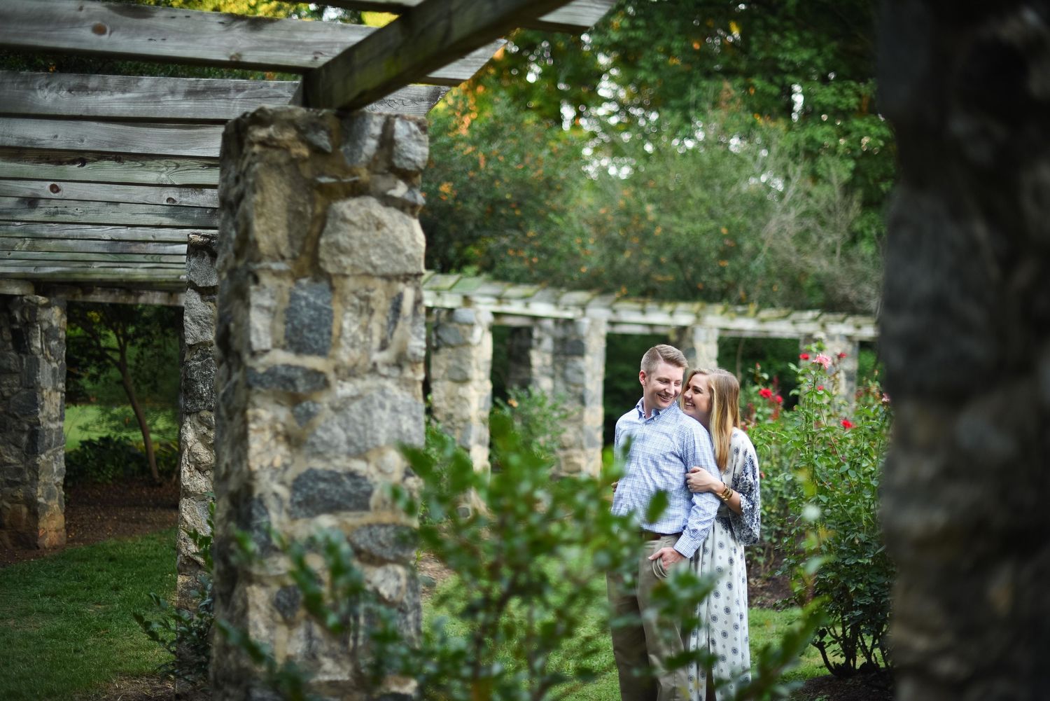 Couple embracing in a lush garden surrounded by stone columns and pergola, romantic outdoor engagement photo.