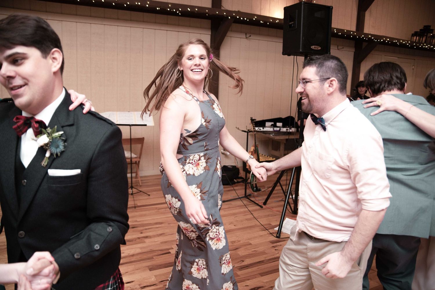 Guests dancing joyfully at a wedding reception in a rustic venue with warm string lights overhead.