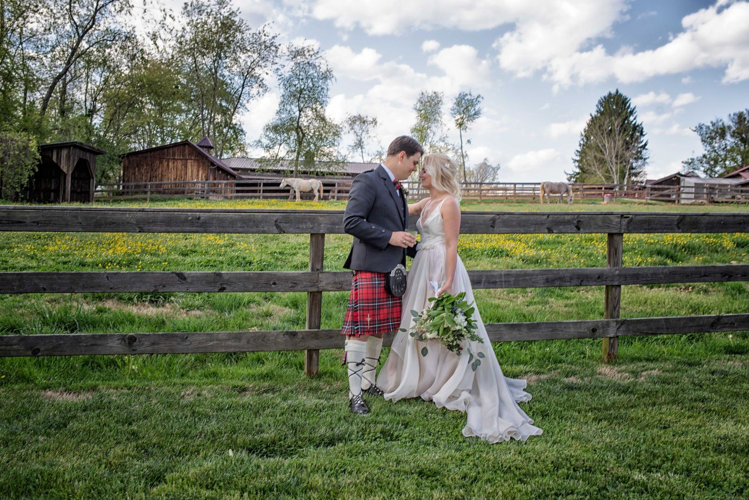 A bride and groom in a red tartan kilt stand by a wooden fence at a rustic farm wedding venue.