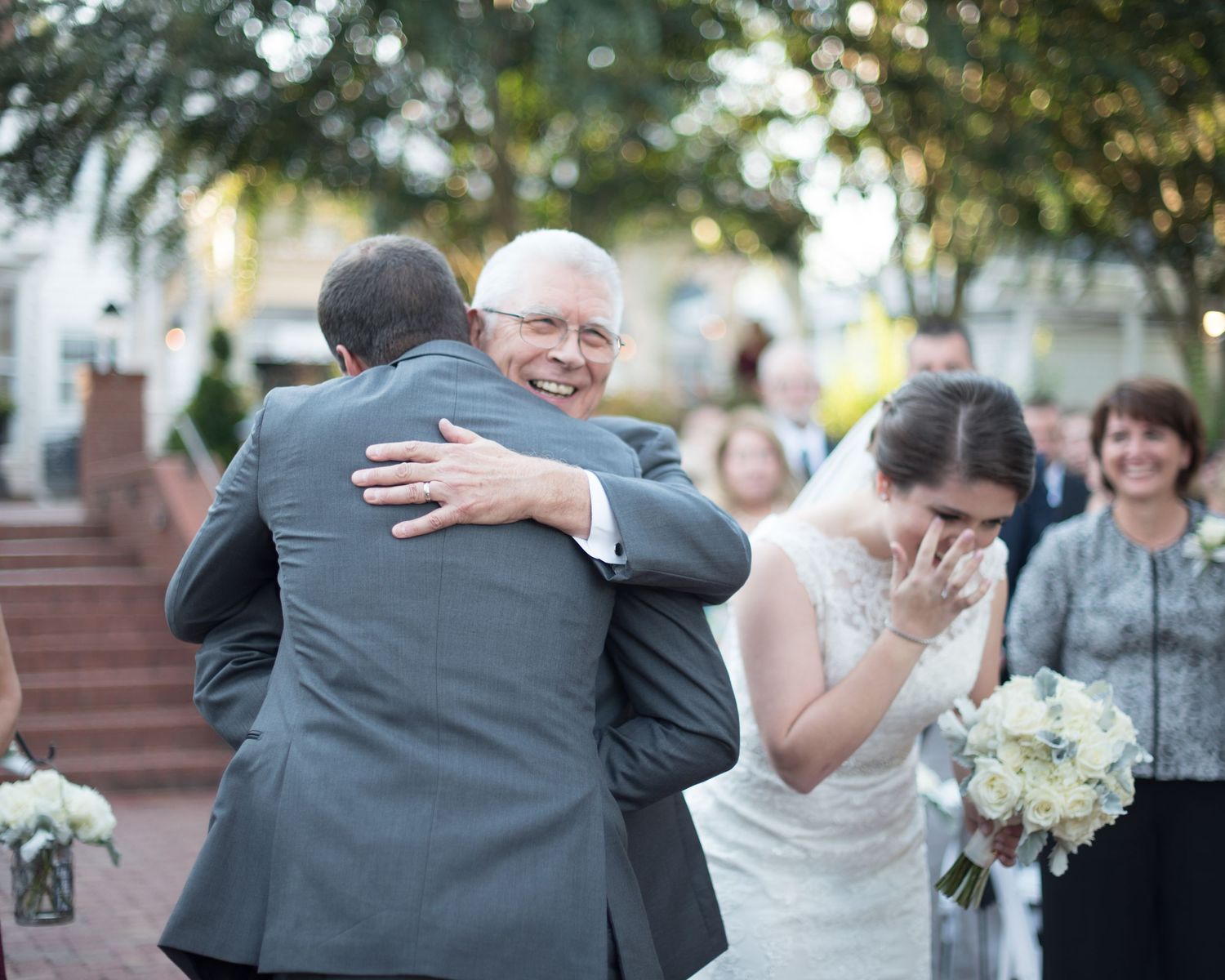 Emotional wedding moment as groom hugs older man while bride wipes tears holding white bouquet.
