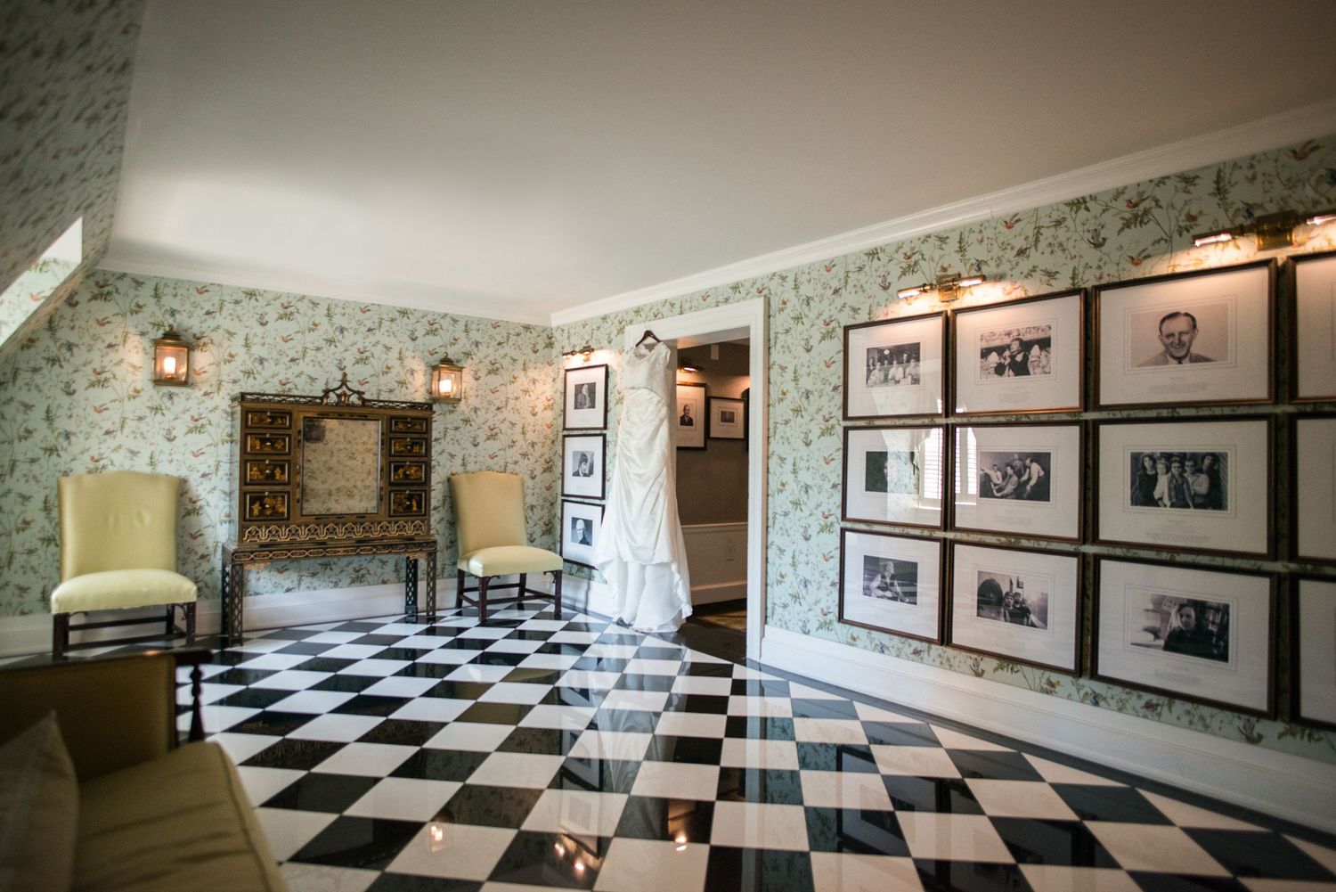 Elegant wedding dress hanging in a vintage room with black and white checkered floors and framed wall photos.