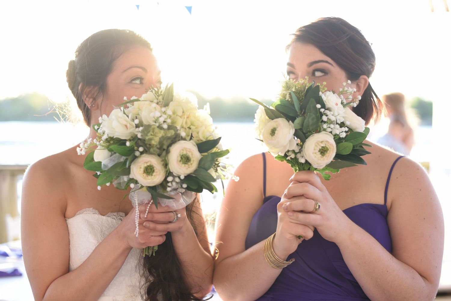 Bride and bridesmaid playfully hiding behind white bouquets at an outdoor wedding ceremony near water.