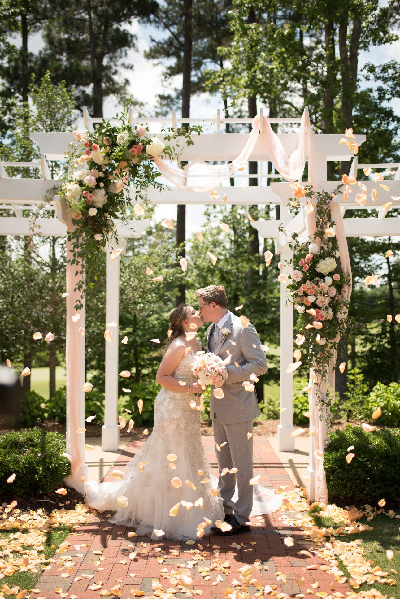 Bride and groom kissing under floral pergola with rose petals falling at outdoor wedding.