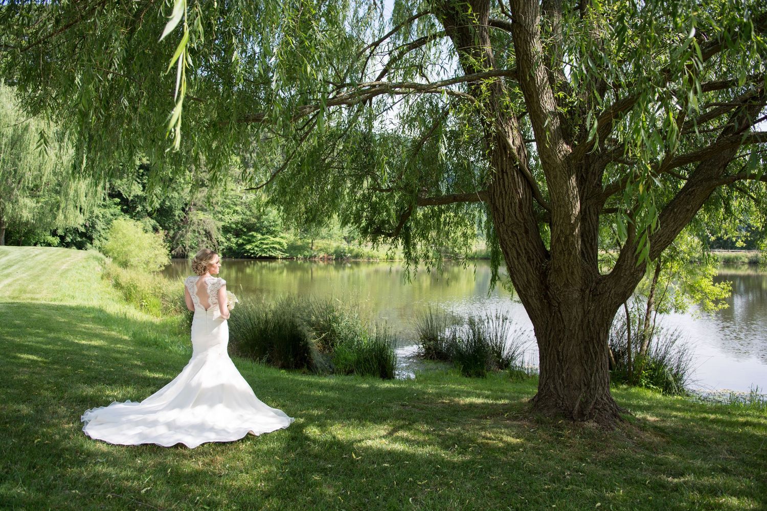 Bride in elegant mermaid gown standing beside willow tree overlooking serene pond.