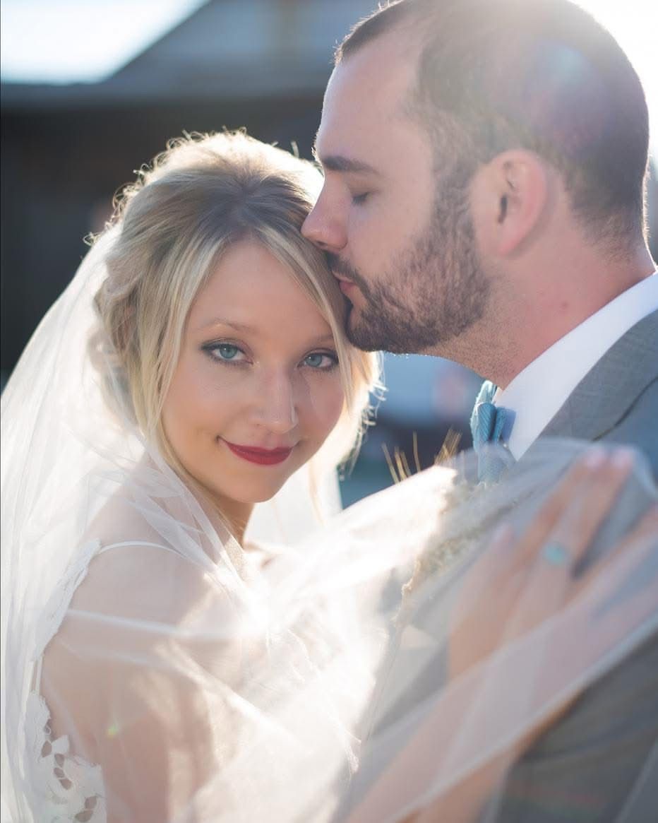 Groom tenderly kissing bride on forehead while she smiles in golden sunlight with veil.