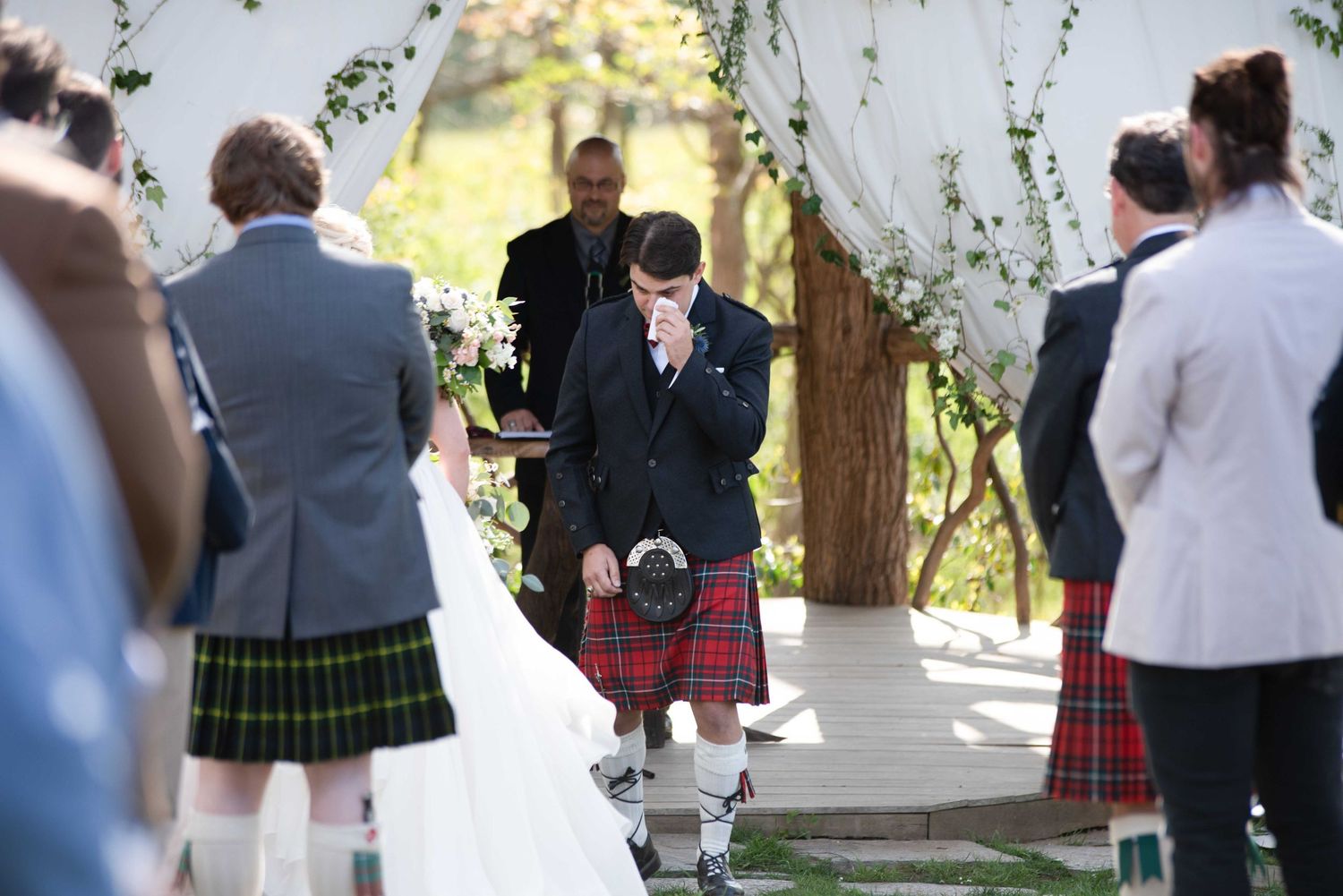 Emotional groom in red tartan kilt crying at outdoor wedding ceremony with guests in kilts and floral arch.