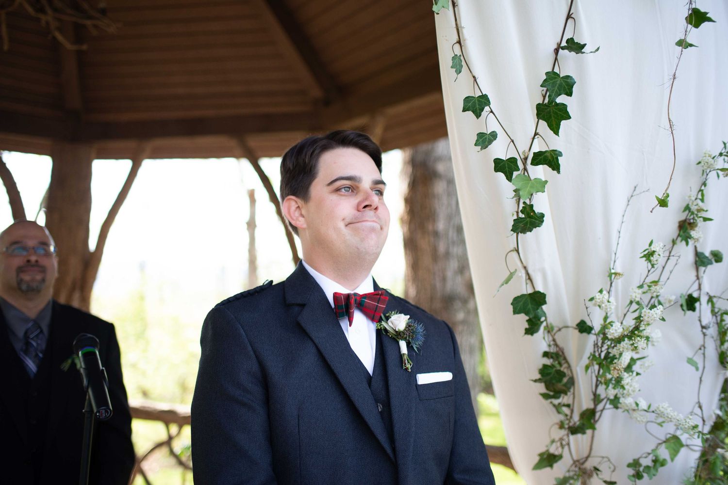 Groom in navy suit and red bow tie stands at outdoor wedding altar decorated with ivy and white draping.