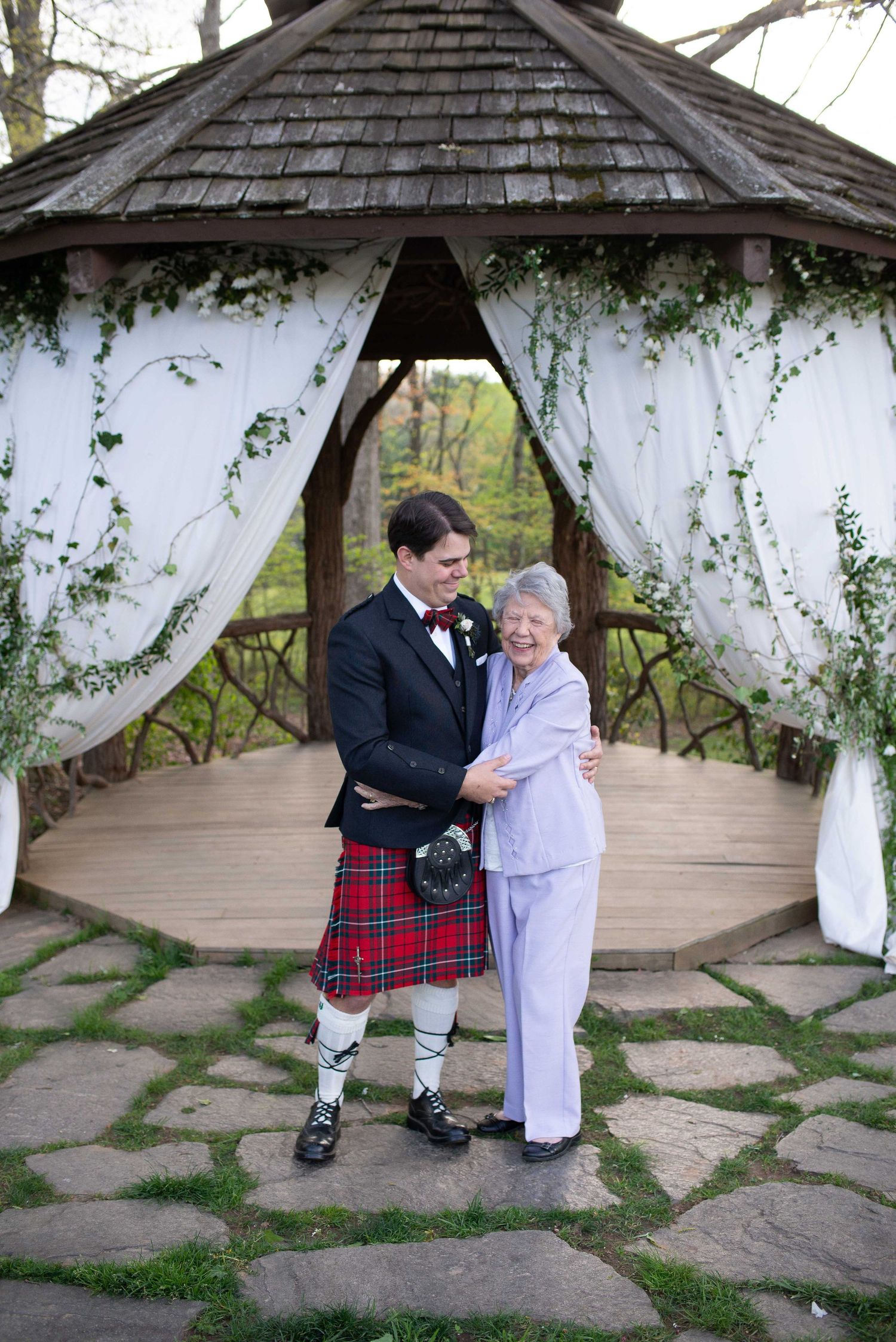 Groom in red tartan kilt poses with elderly woman before decorated gazebo at outdoor wedding ceremony.
