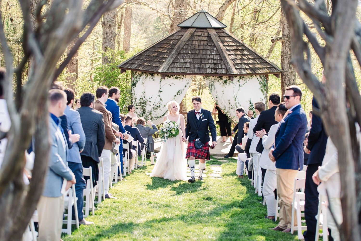 Outdoor wedding ceremony under a gazebo with guests seated on white chairs along a garden aisle.