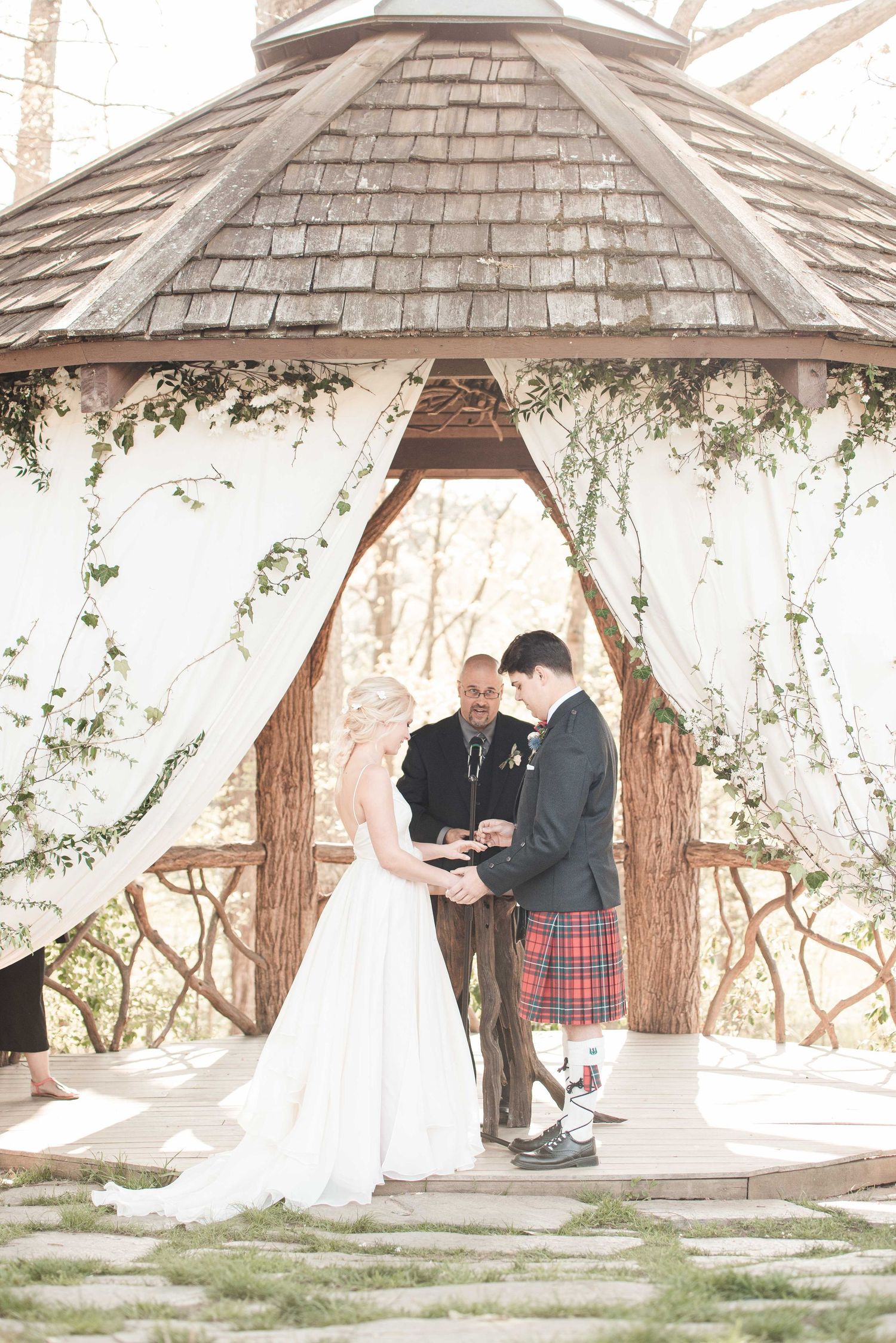 Bride and groom exchange vows under a rustic gazebo draped in white fabric and greenery, groom wearing a red tartan kilt.