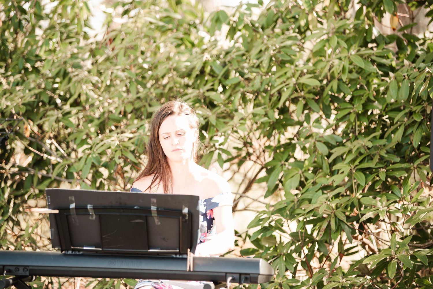 Woman playing keyboard piano outdoors surrounded by lush green trees on a sunny day.
