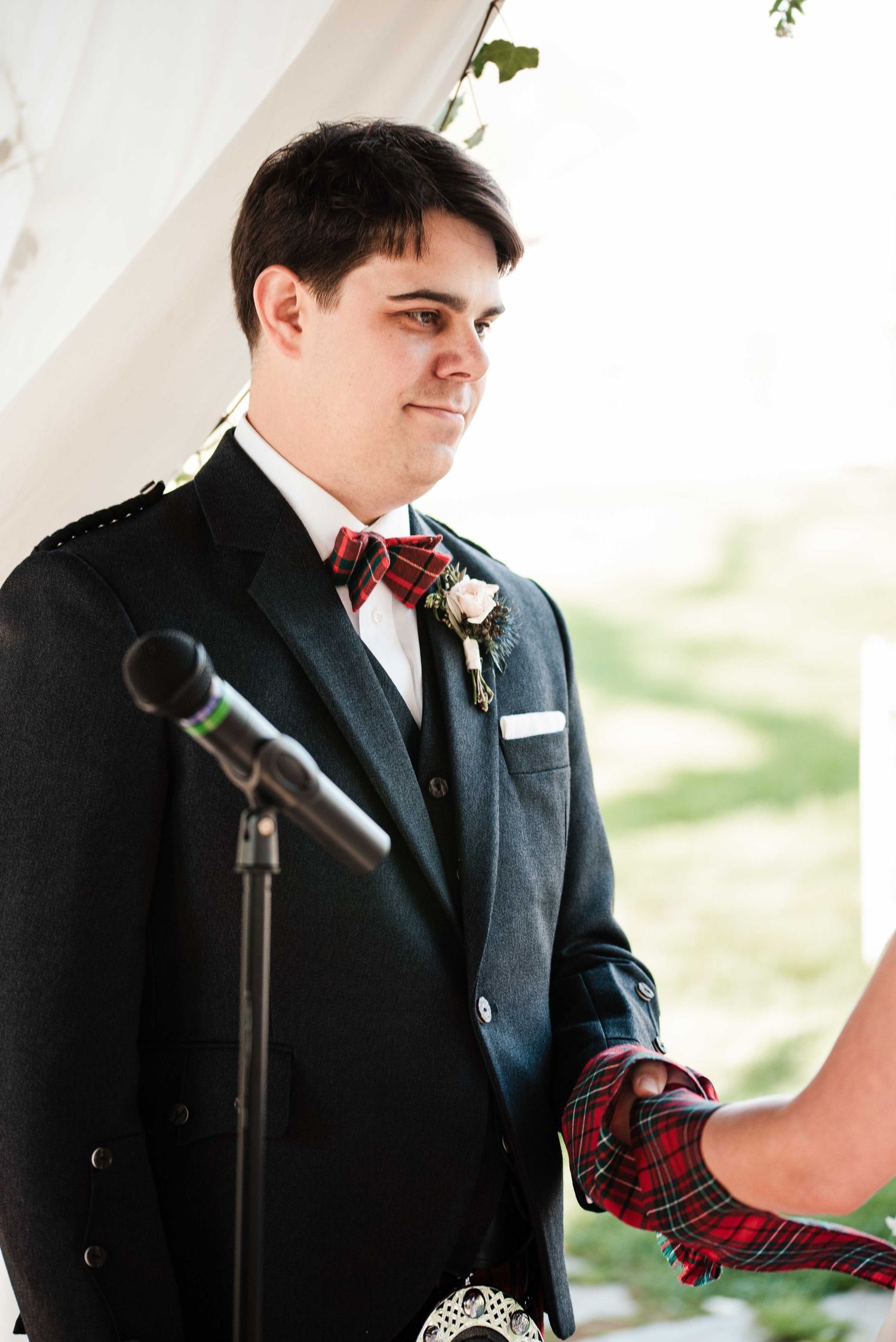 Groom in black suit with tartan bow tie and boutonniere holds hands at outdoor wedding ceremony.