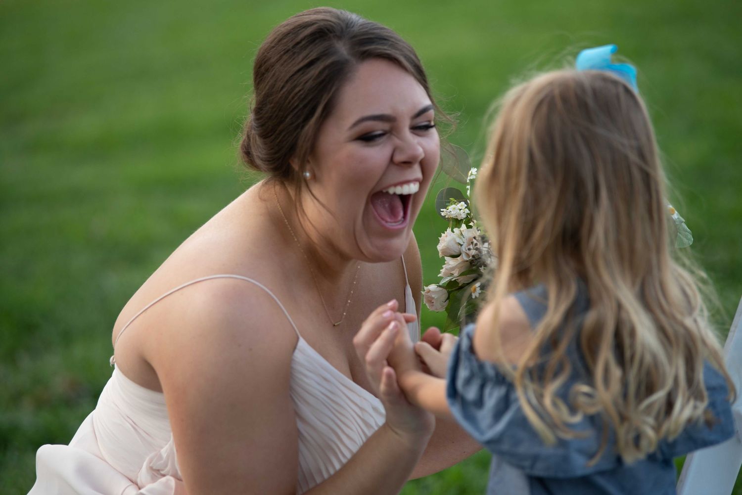 A bride laughs joyfully as a young flower girl presents her with a small bouquet of white flowers outdoors.