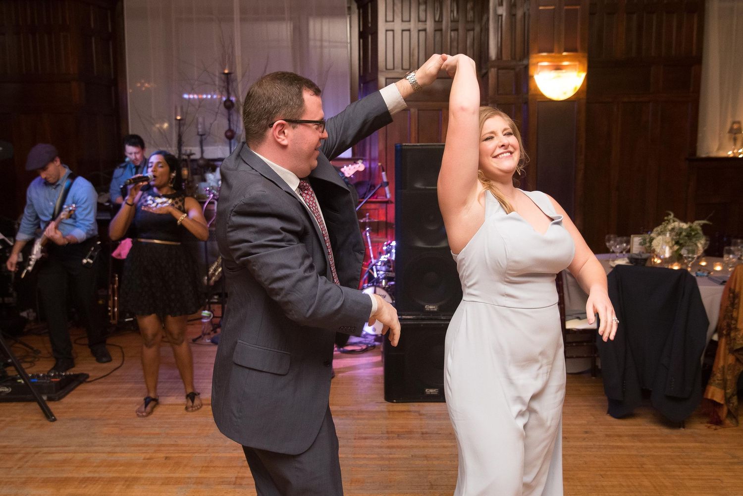 Couple dancing at wedding reception, man spinning woman in gray dress on dance floor with live band performing in background.