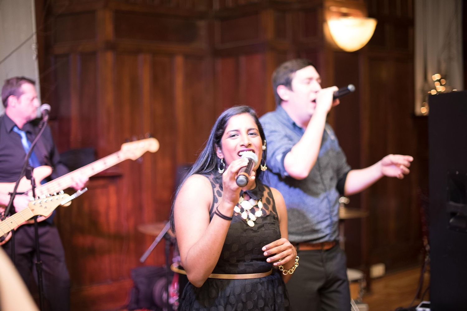 Live band performance with female vocalist singing on stage, guitarist and male singer performing in background.