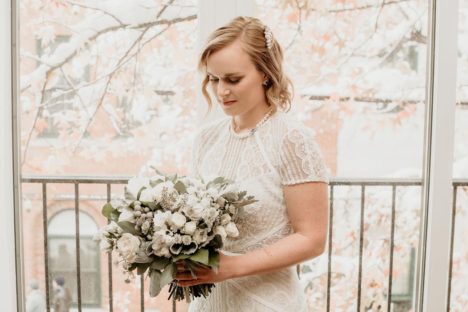 Bride in beaded white gown holding elegant white and greenery bouquet, standing on balcony with blooming trees in background.