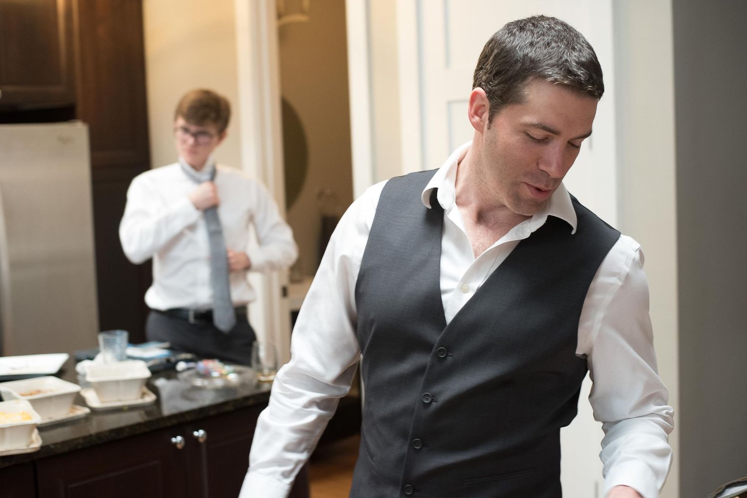 Two men in formal attire adjusting their ties and vest in a well-lit room, preparing for a special occasion.
