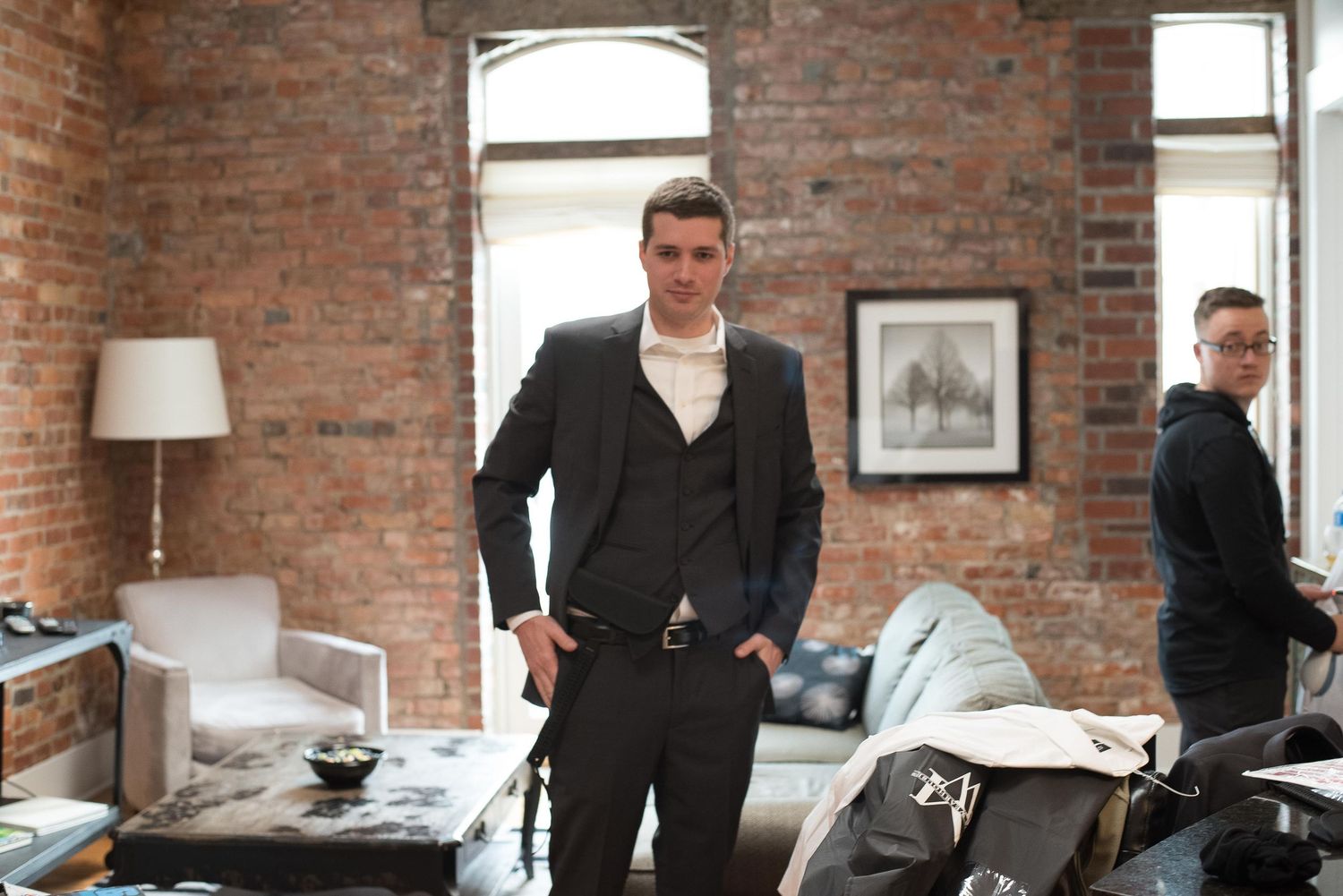 Man in dark suit stands confidently in industrial brick loft apartment with contemporary furnishings.