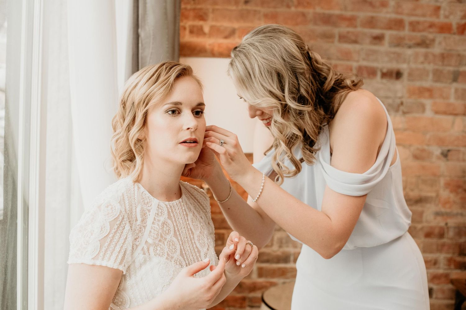 Bridesmaid helping bride put on earrings near window with brick wall backdrop on wedding day.