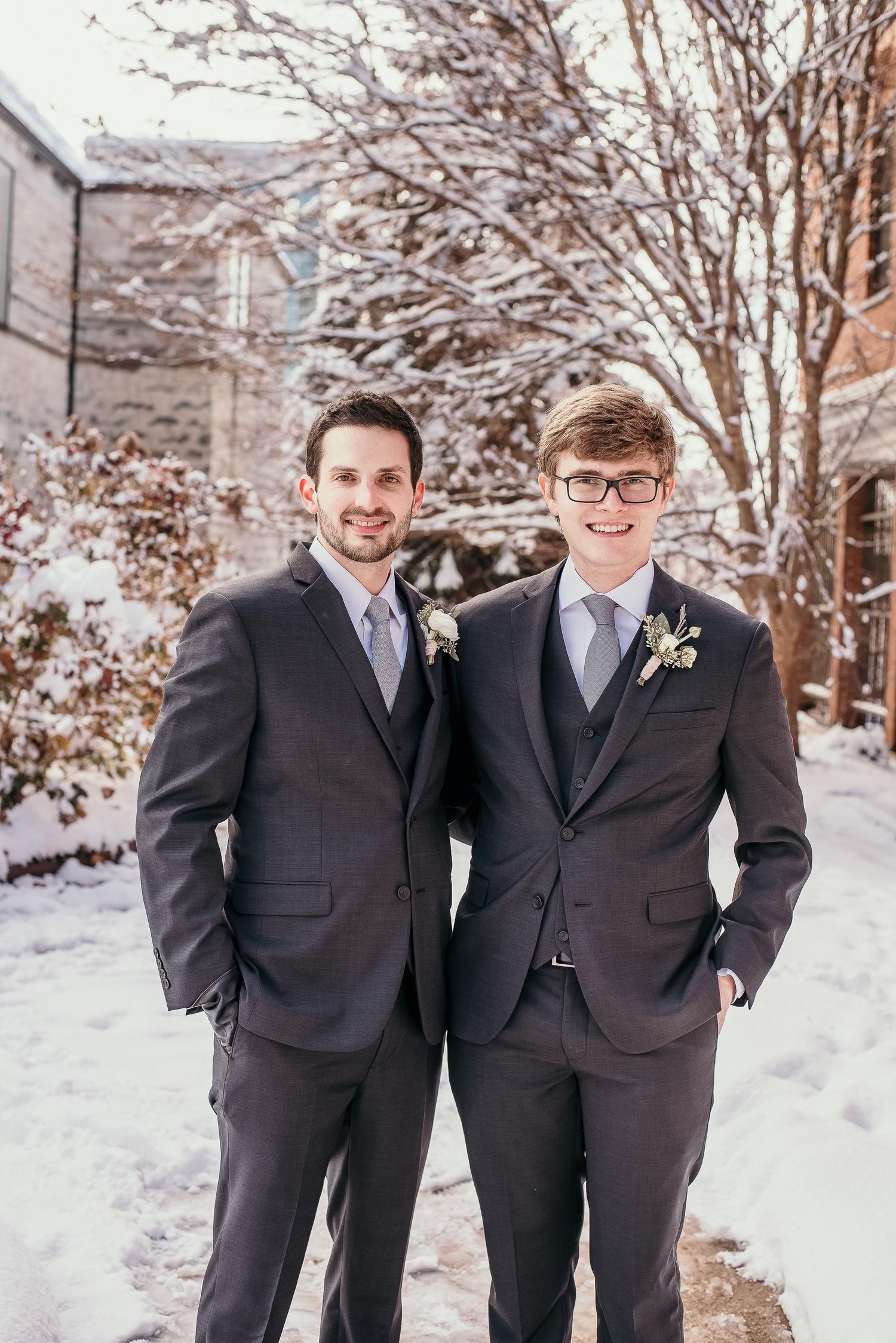 Two men in matching dark suits with boutonnieres pose outdoors in a snowy winter setting.