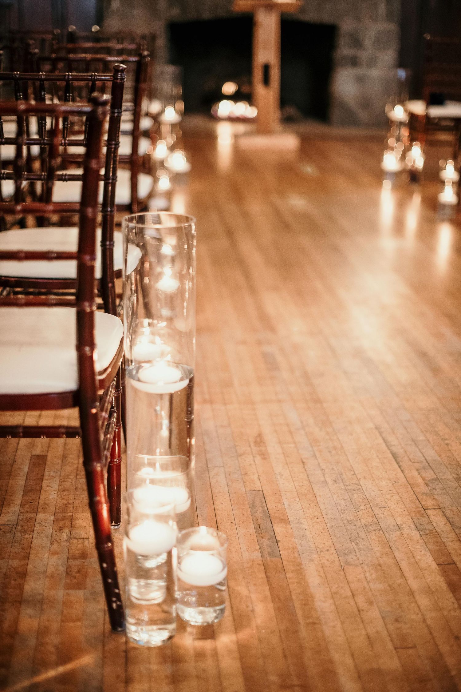 Floating candles in glass cylinders line a wooden aisle at an elegant indoor wedding ceremony.